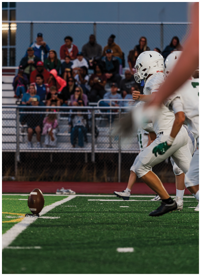Several people in football uniforms are on a field with a football placed upright on a tee, and a crowd is seated in bleachers in the background. Description generated by AI