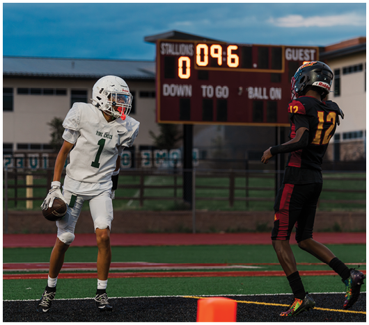A person in white football gear holding a football stands on a field near the end zone while another person in black and red football gear approaches, with a scoreboard showing the score and time in the background. Description generated by AI