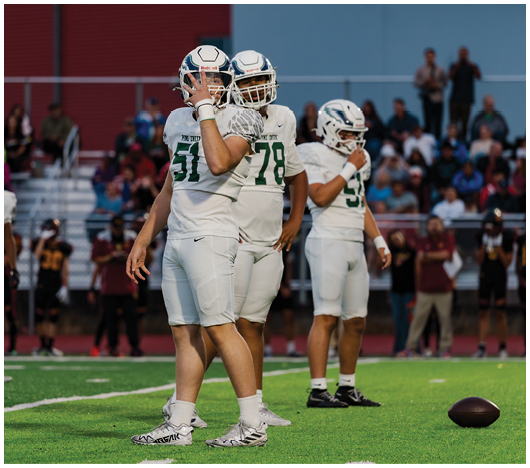 Three people in white football uniforms and helmets stand on a green field with a football on the ground nearby and a crowd in the background. Description generated by AI