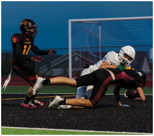 Two people in football gear are engaged in a tackle on a field near a goalpost during a game. Description generated by AI