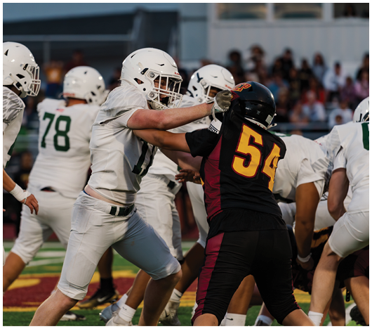 Two football players in white and black uniforms are engaged in a physical block during a game on a field with other players and spectators in the background. Description generated by AI