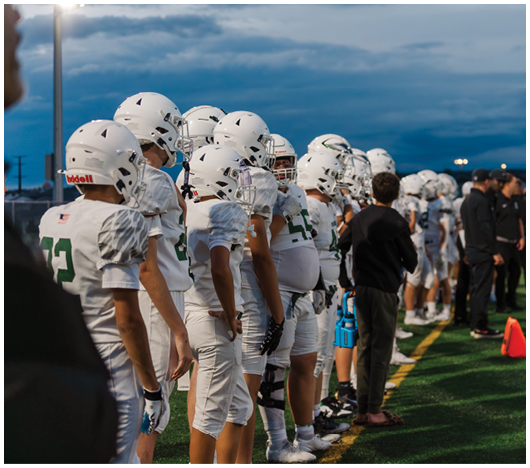 A line of people wearing white football uniforms and helmets stand on the sidelines of a field under a cloudy evening sky. Description generated by AI