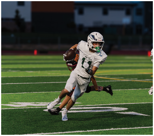A person in white football gear is running with the ball while two other players in white gear and one player in dark gear attempt to catch or block them on a green football field. Description generated by AI