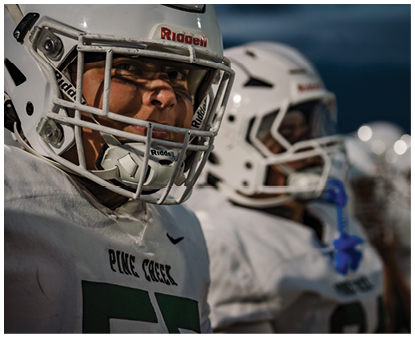 People wearing white football helmets and jerseys stand in a line, with one person in the foreground showing a focused expression. Description generated by AI