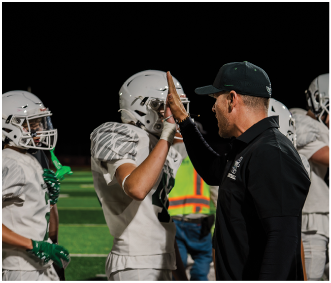 A person in a black shirt is giving a fist bump to a person in white football gear on a field at night. Description generated by AI