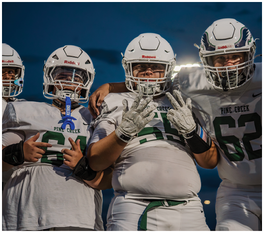Four people in football uniforms and helmets stand closely together outdoors at dusk, with one person in the center making a hand gesture. Description generated by AI