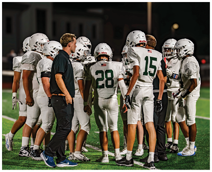 A group of people wearing sports uniforms and cleats are standing together on a grassy field, likely during a team huddle or discussion. Description generated by AI