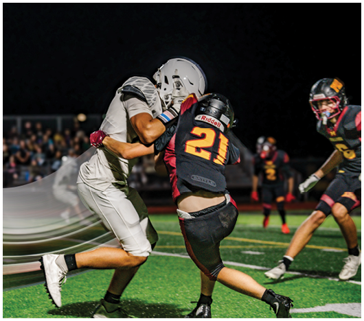 Two people in football uniforms are engaged in a tackle on a field with other players and spectators visible in the background. Description generated by AI