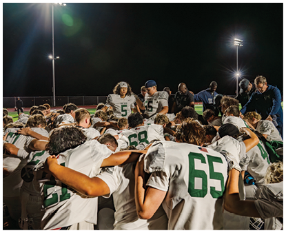 A group of football players in white uniforms with green numbers are huddled together on a field at night under bright stadium lights. Description generated by AI