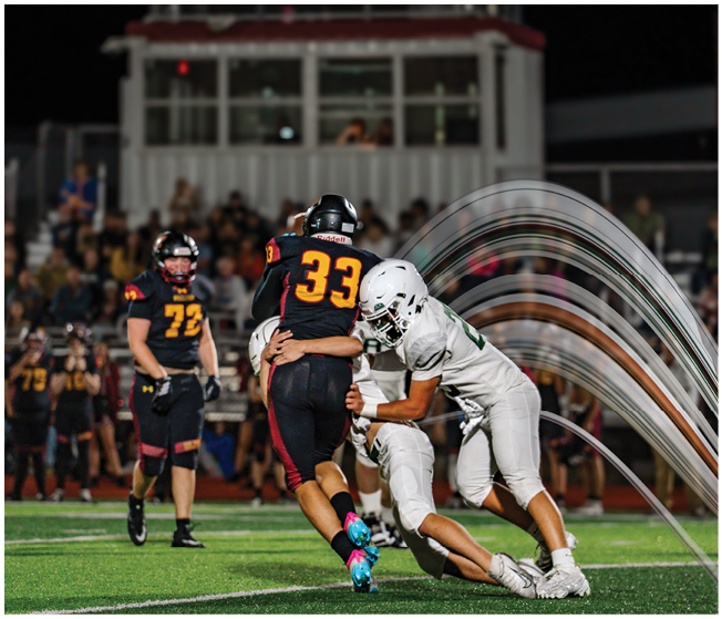 Two people in football uniforms are engaged in a tackle on a field while another person in uniform watches nearby with a crowd in the background. Description generated by AI