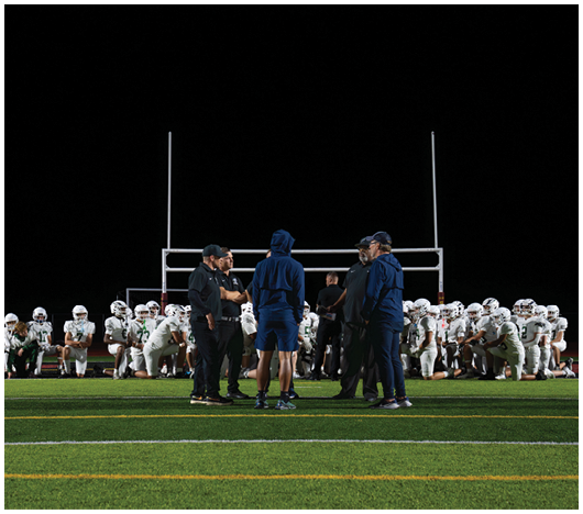 A group of people in sportswear stands on a football field with two teams of players in white uniforms kneeling behind them near the goalposts at night. Description generated by AI