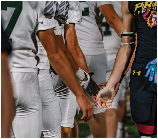People in football uniforms are standing in a line reaching out to shake hands with a person wearing a black sleeveless shirt and blue gloves. Description generated by AI