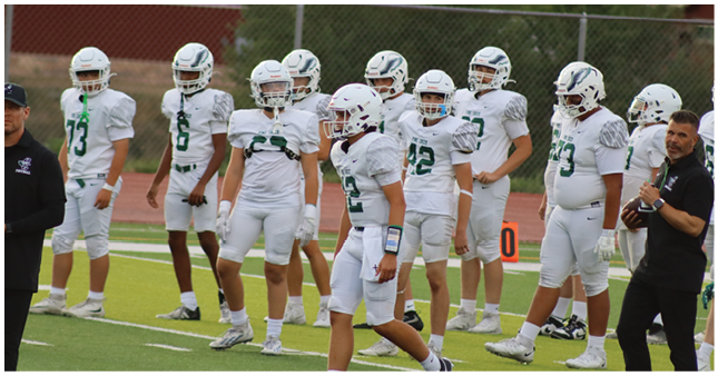 A group of people wearing white football uniforms and helmets stand on a field near a chain-link fence with two adults in black shirts nearby. Description generated by AI
