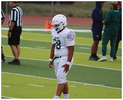 A person in a white football uniform and helmet stands on a green field with two referees and two other people in the background. Description generated by AI
