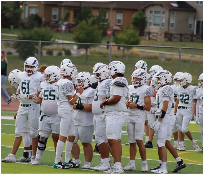 A group of people wearing white football uniforms and helmets are gathered on a field while a person in dark athletic clothing stands nearby. Description generated by AI