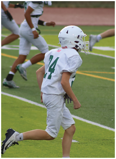 A person wearing a white football uniform and helmet is running on a green field with other players in the background. Description generated by AI