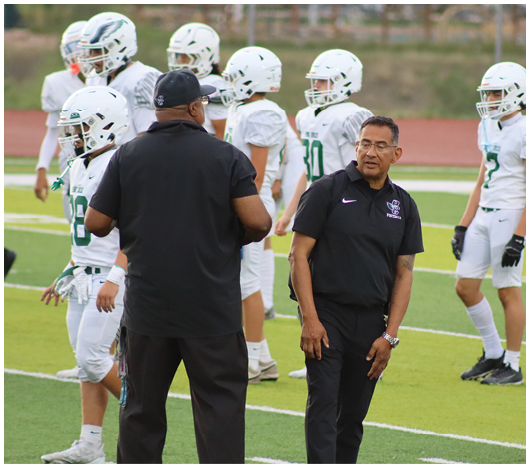 A group of people in white football uniforms and helmets stand on a field while two people in black shirts and pants appear to be coaching or instructing them. Description generated by AI