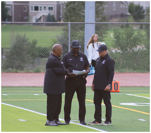Three adults wearing black sportswear stand on a football field discussing a document while a person in a white jacket stands in the background near a chain-link fence. Description generated by AI