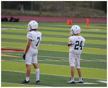 Two children wearing white football uniforms and helmets stand on a green field with yard lines and markers. Description generated by AI
