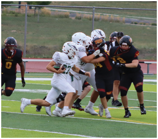 A group of people wearing football uniforms are actively engaged in a play on a green field with a chain-link fence and grassy area in the background. Description generated by AI