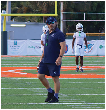 An adult wearing a navy blue outfit and cap stands on a football field with three people in white sports uniforms stretching behind them and a scoreboard showing zero scores in the background. Description generated by AI