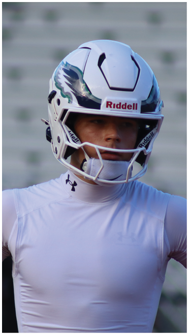 A person wearing a white football helmet and a white athletic shirt stands on a field with blurred bleachers in the background. Description generated by AI