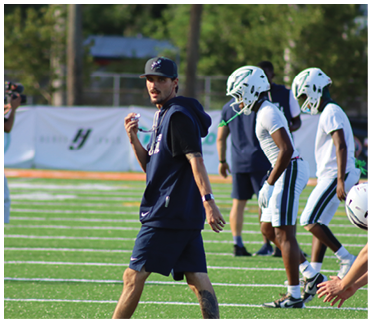 A person wearing a cap and sportswear is holding a whistle on a football field while two people in football helmets and gear stand nearby and another person films in the background. Description generated by AI