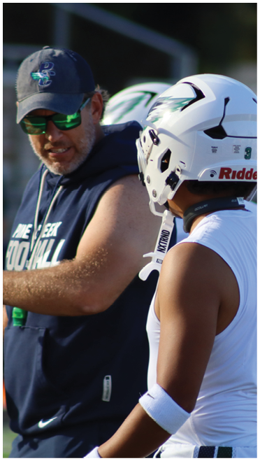 An adult wearing a cap and sunglasses is talking to a person in a football helmet and sleeveless athletic shirt on a sports field. Description generated by AI