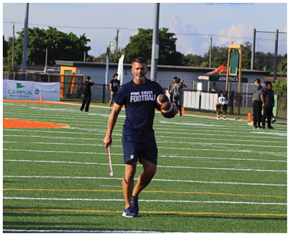 An adult wearing a navy blue Pine Creek Football shirt and shorts is walking on a football field holding a football and a flag while other people stand or walk in the background. Description generated by AI