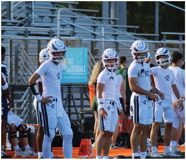 Four people wearing white football uniforms and helmets stand on the sidelines of a football field with bleachers and other people in the background. Description generated by AI