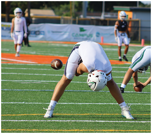 A person in white sports gear is bending over on a football field while holding a football, with other players standing in the background. Description generated by AI