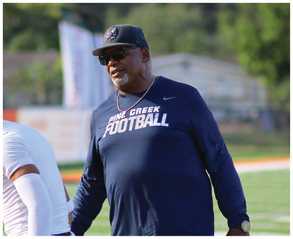 A person wearing a navy blue long-sleeve shirt with “Pine Creek Football" printed on it stands on a sports field with other people in athletic clothing in the background. Description generated by AI