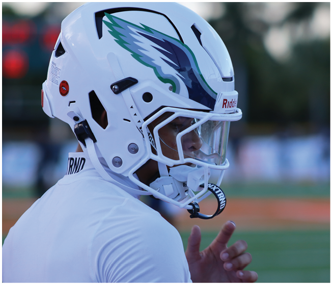 A person wearing a white football helmet with a green and blue eagle logo is standing on a field, appearing focused and ready for action. Description generated by AI