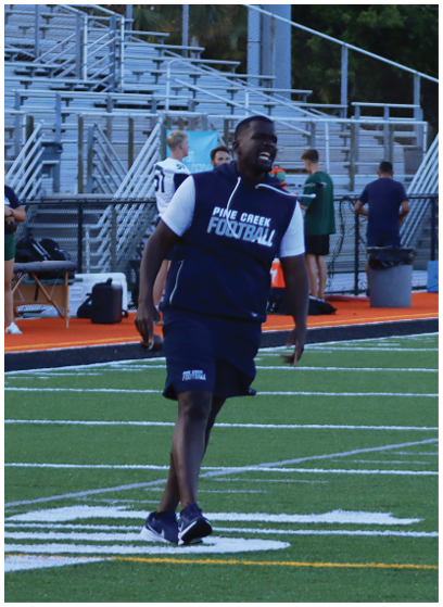 A person wearing a Pine Creek Football sleeveless hoodie and shorts is walking on a football field with bleachers and other people in the background. Description generated by AI