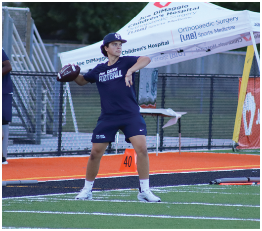 A person wearing a sports outfit is preparing to throw a football on a field with yard markers and a tent in the background displaying hospital sponsorship. Description generated by AI