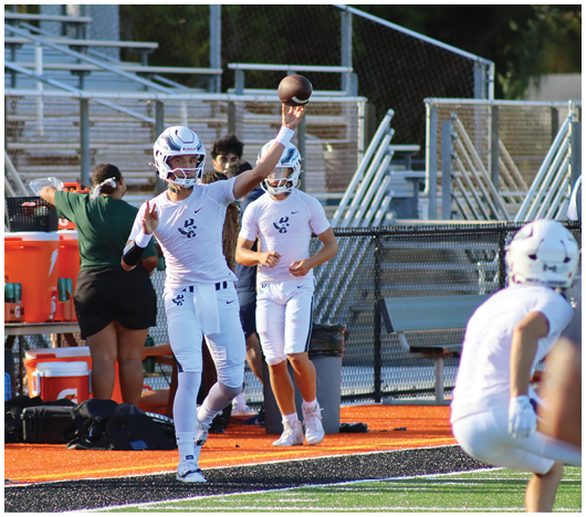 A person in a white football uniform is throwing a football while two other people in similar uniforms and one person in casual clothing stand nearby on the sidelines of a football field. Description generated by AI