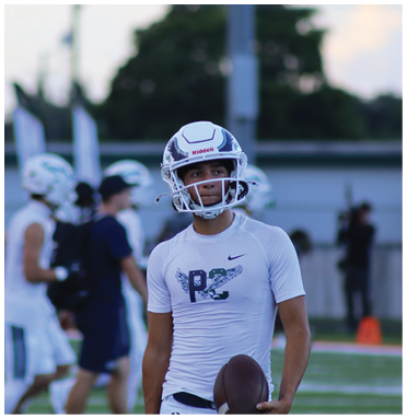 A person wearing a white football helmet and white athletic shirt is holding a football on a field with other people in similar sports attire in the background. Description generated by AI