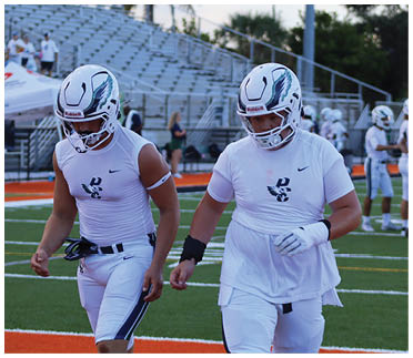 Two people wearing white football helmets and athletic gear walk on a football field with other players and empty bleachers in the background. Description generated by AI