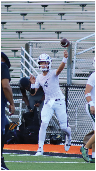 A person in white athletic gear and helmet is throwing a football on a field with empty bleachers in the background. Description generated by AI