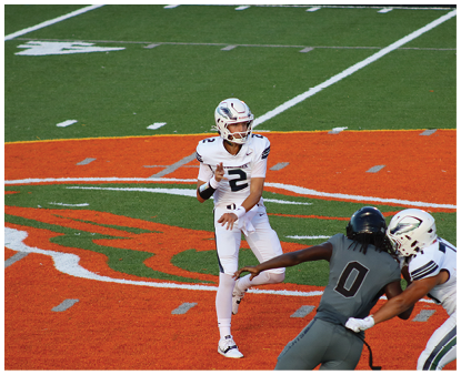 A person in a white football uniform is preparing to throw a football while two people in dark and white uniforms are approaching on an orange and green field. Description generated by AI