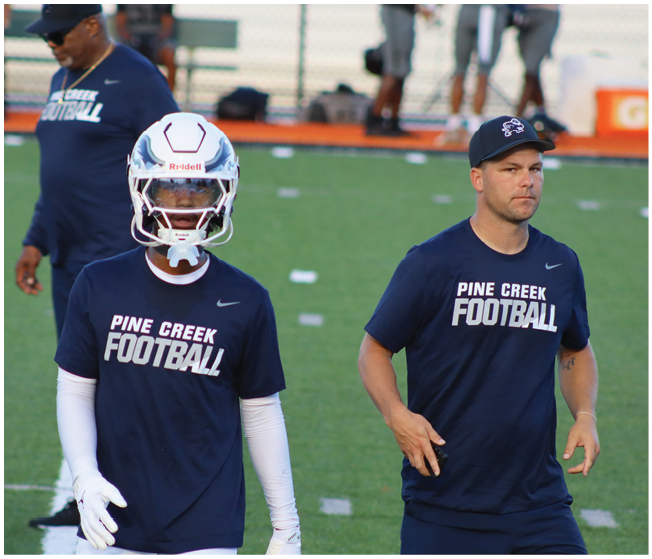 People wearing Pine Creek Football shirts and helmets are standing on a football field during practice or warm-up. Description generated by AI