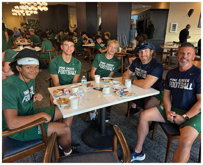 A group of people wearing matching green and blue Pine Creek Football shirts are sitting around a table in a dining area, smiling and eating a meal together. Description generated by AI