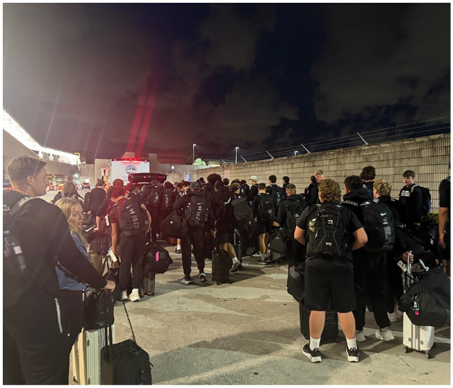 A large group of people dressed in black clothing and carrying backpacks and luggage are standing in line outdoors at night near a concrete wall and a building with bright lights. Description generated by AI