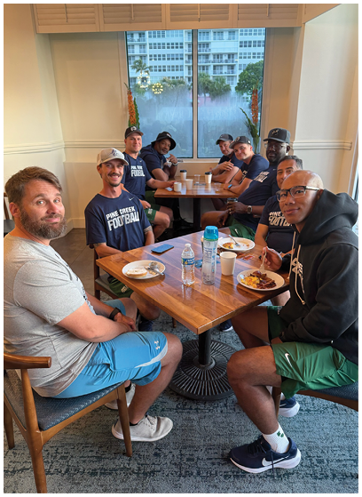 A group of adults wearing casual sportswear sit around two wooden tables in a dining area with plates of food and drinks, smiling and engaging with each other. Description generated by AI