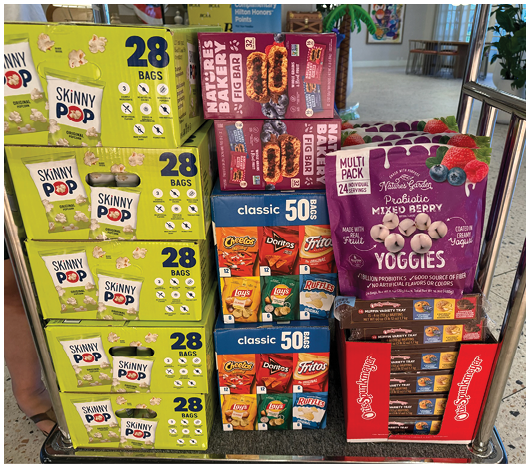 A display cart holds multiple boxes of Skinny Pop popcorn, Nature's Bakery fig bars, a variety pack of classic snack chips, and a multi-pack of probiotic mixed berry yogis. Description generated by AI