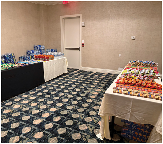 Two tables covered with white and black tablecloths display an assortment of snacks and bottled drinks in a carpeted room with beige walls and a closed door. Description generated by AI