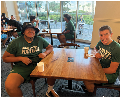 Two people wearing green sports uniforms sit at a wooden table in a casual dining area, smiling and holding drinks. Description generated by AI