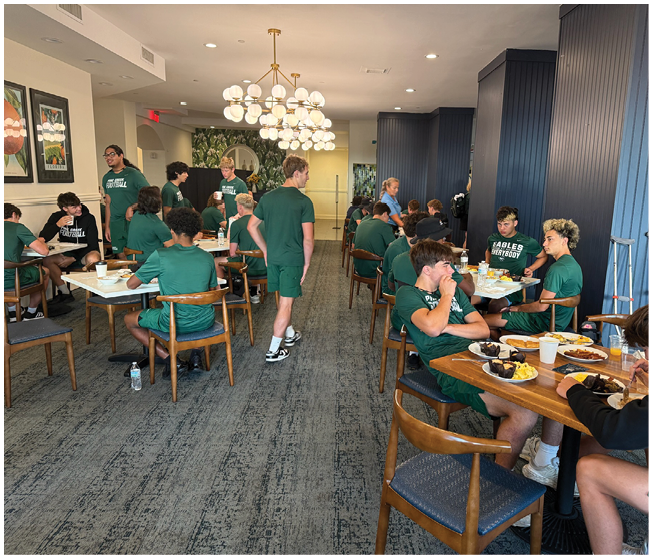 A group of people wearing matching green shirts are sitting and standing around tables in a well-lit dining area, eating and socializing. Description generated by AI