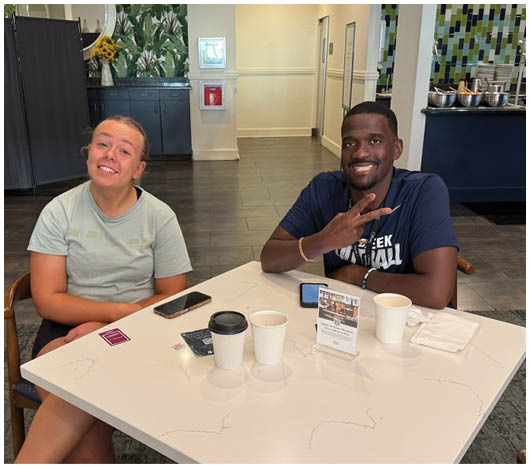 Two adults sit at a table in a casual indoor setting smiling, with cups and phones on the table. Description generated by AI