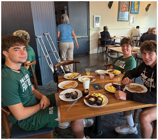 Three young people wearing matching sports uniforms are sitting at a table in a casual dining setting, smiling and eating breakfast with plates of scrambled eggs, waffles, and muffins. Description generated by AI